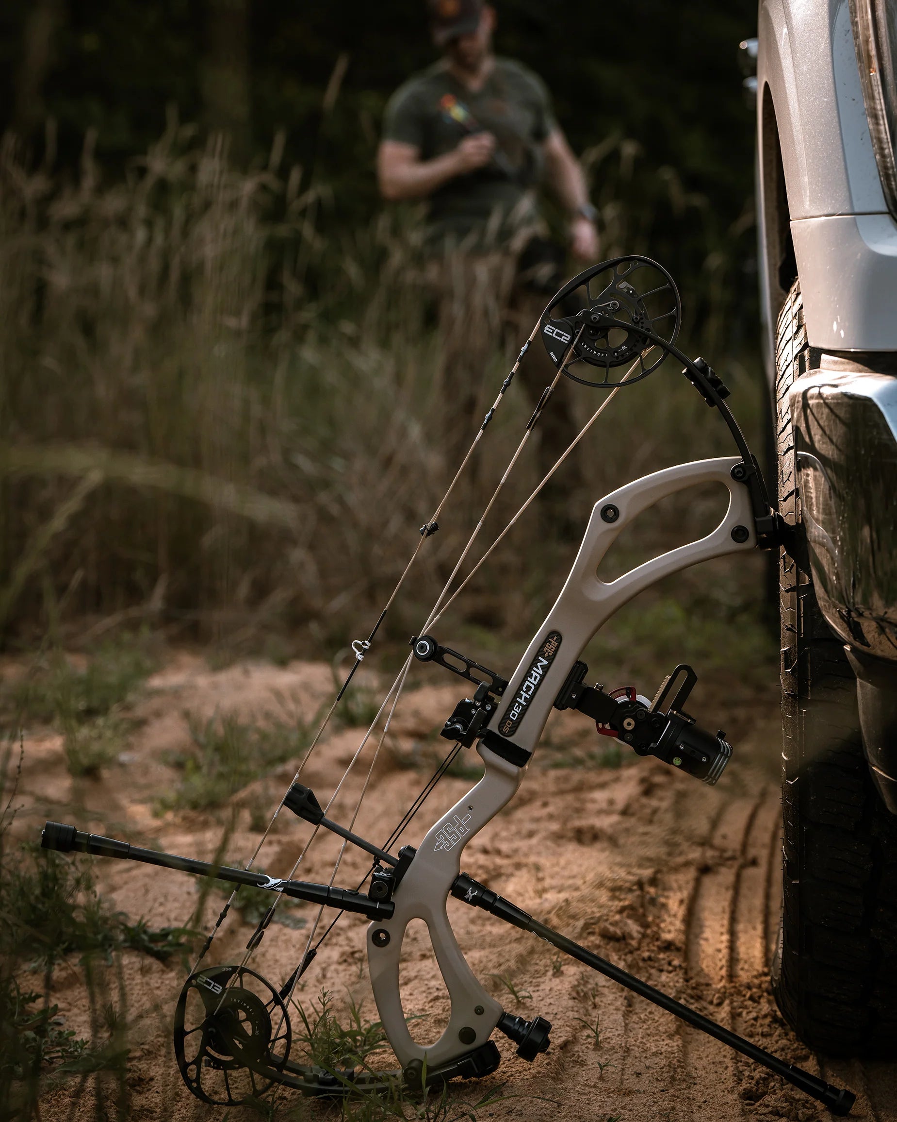 Compound bow attached to a vehicle in a natural setting with a person in the background.