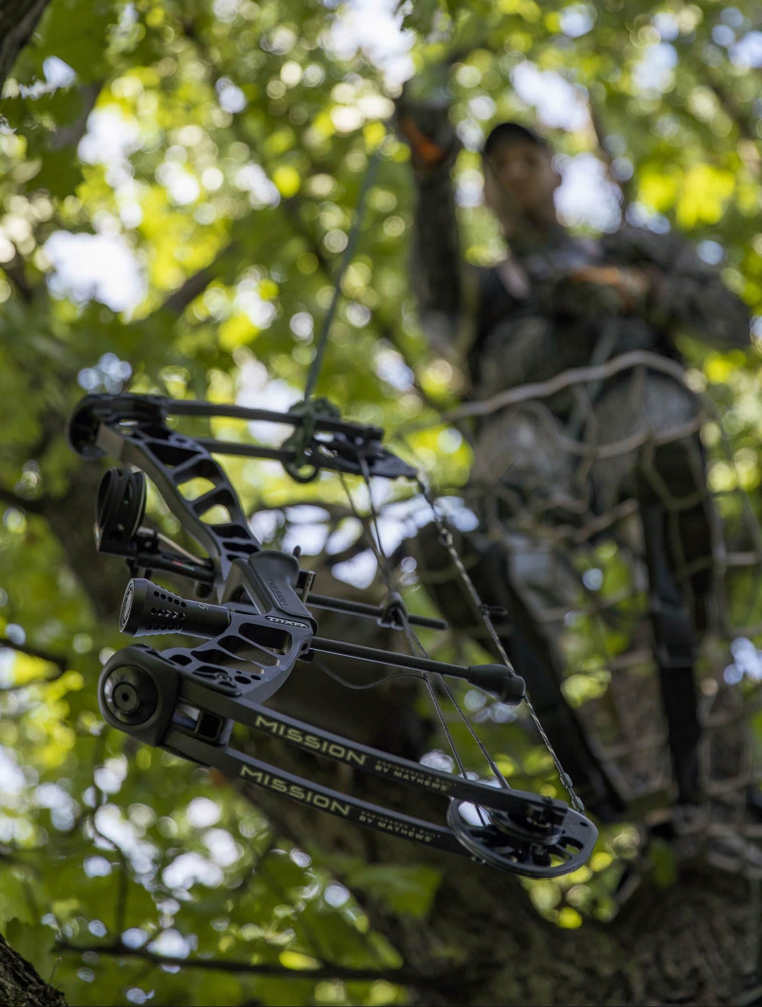 Person in camouflage standing in a tree with a crossbow, surrounded by green foliage.