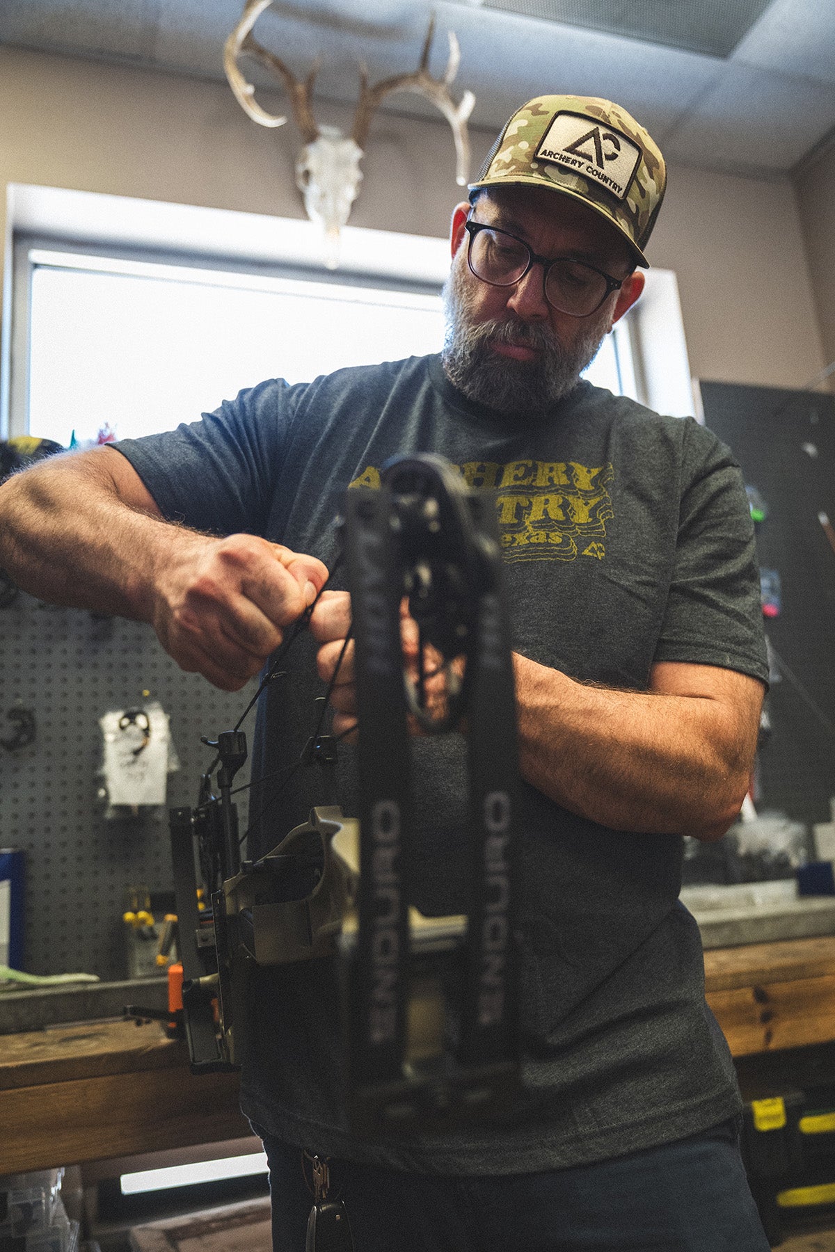 An archer wearing a branded Archery Country cap and tee while prepping gear inside the Austin pro shop.